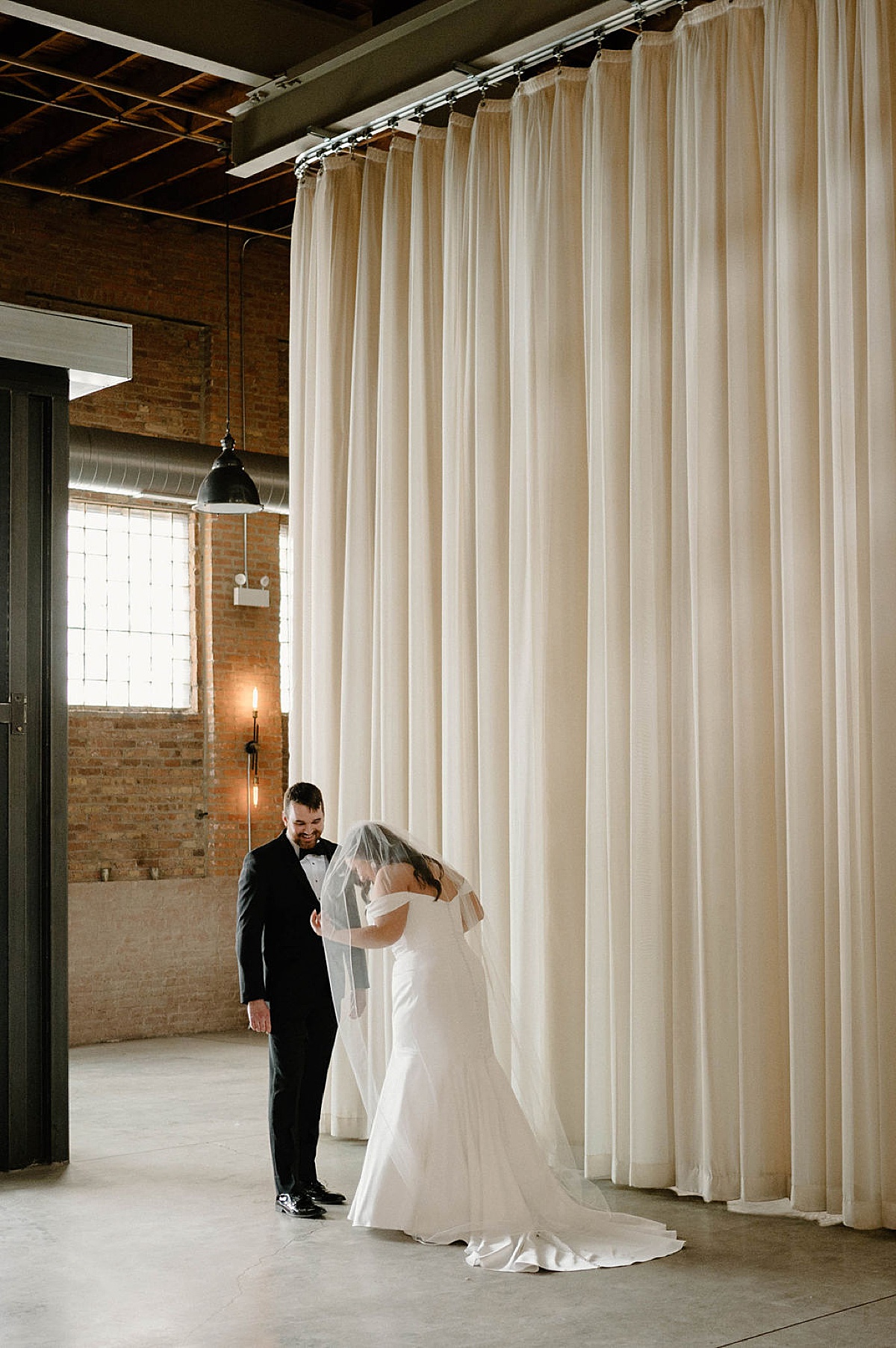 groom smiles as bride adjusts elegant gown and veil during first look shot by Chicago wedding photographer
