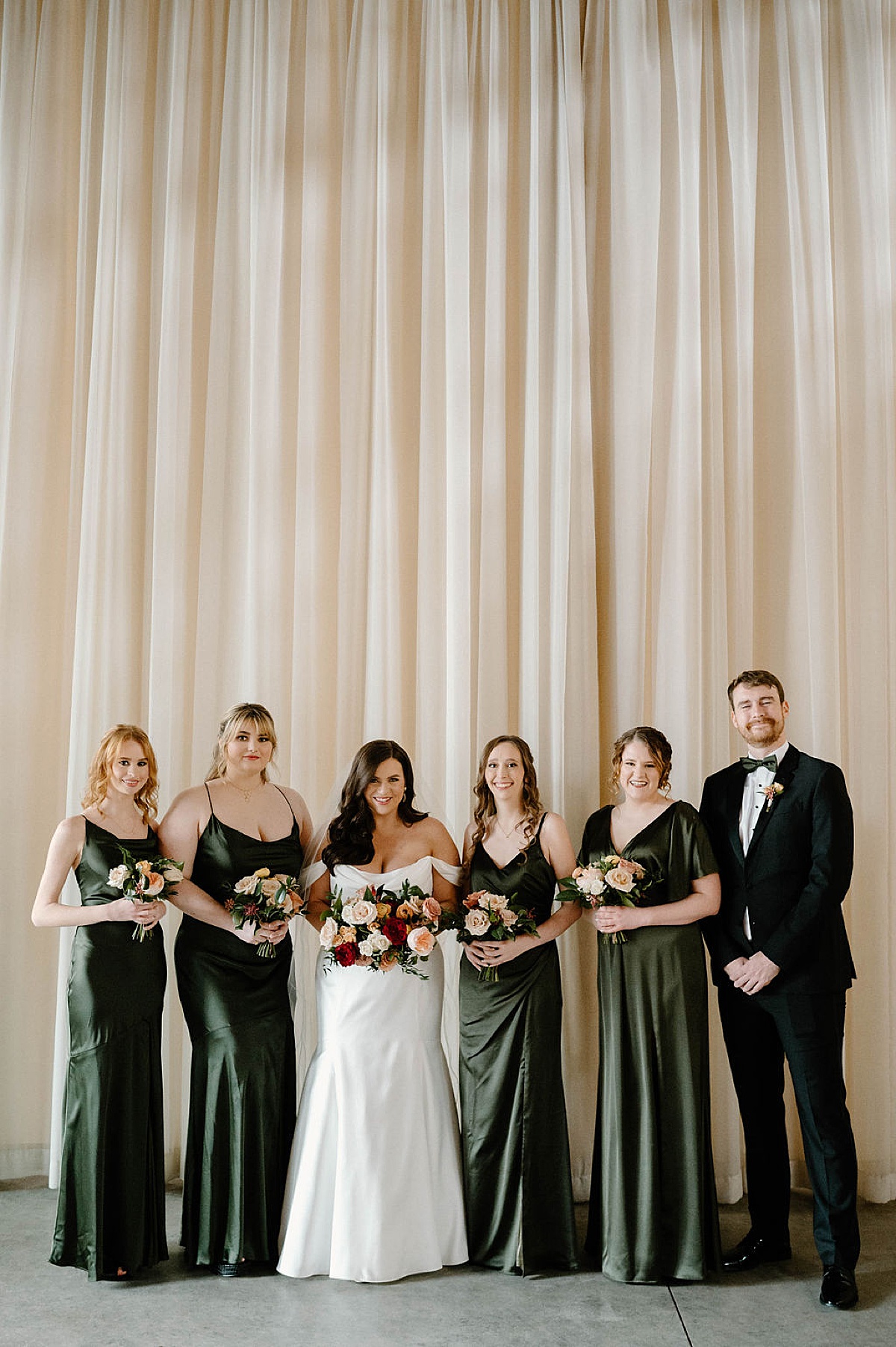 bridesmaids in olive gowns pose with bride holding pink red and white roses during portraits shot by Chicago wedding photographer