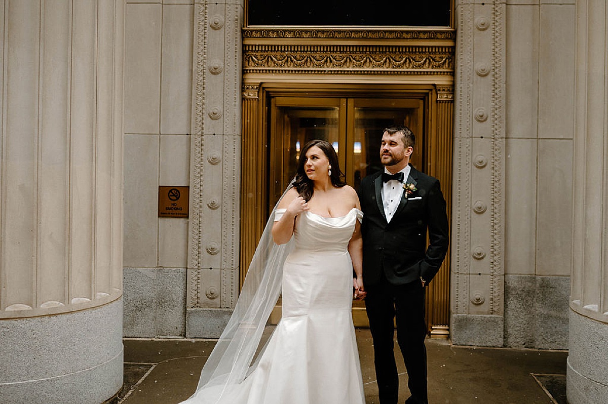 bride in snatched wedding gown with draped neckline poses with dapper groom outside building with gold doors  shot by Indigo Lace Collective