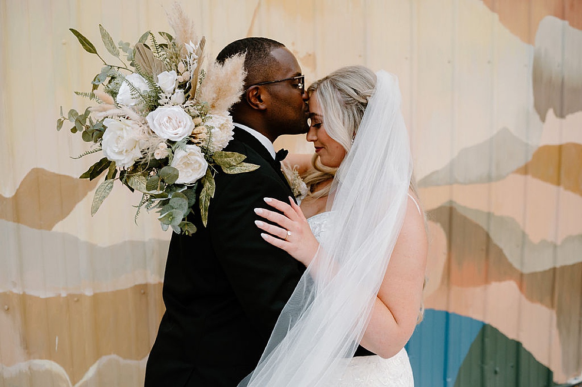 groom kisses bride on the forehead as she holds a boho bouquet of roses and pampas grass after sunny Paper Mills ceremony