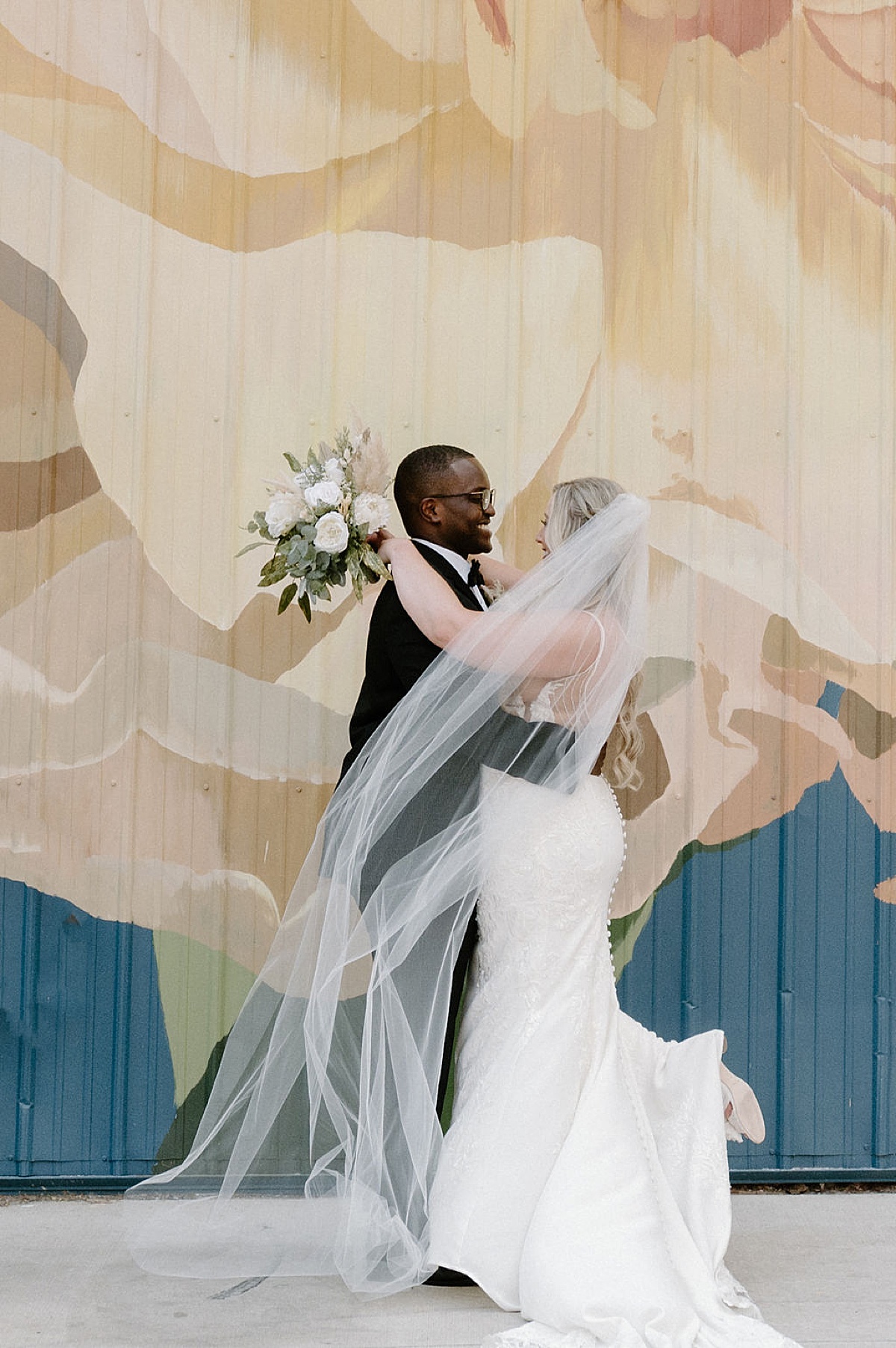 smiling groom embraces bride in elegant lace and satin gown as they pose in front of colorful mural after sunny Paper Mills ceremony