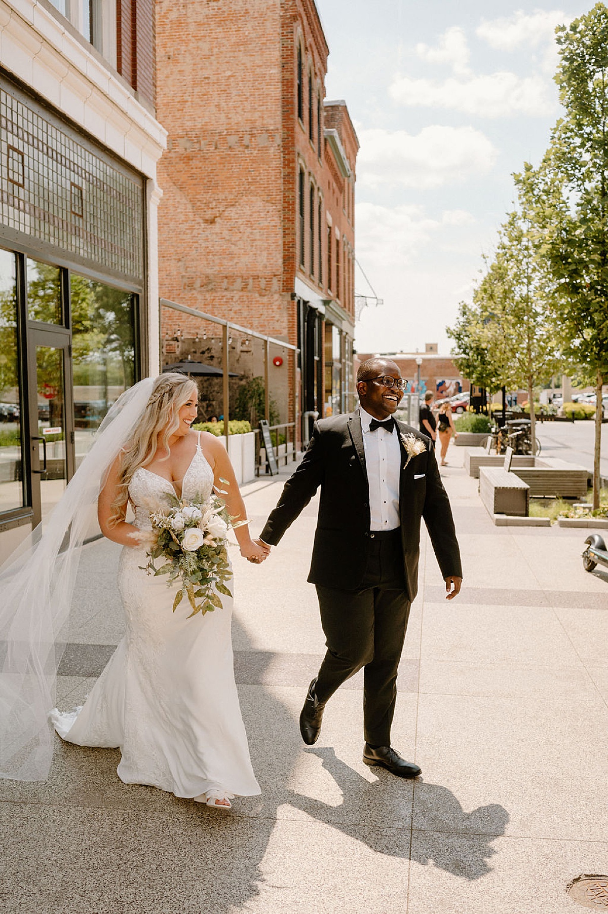cute bride and groom in classy boho evening attire walk along sunny sidewalk after ceremony shot by Indigo Lace Collective