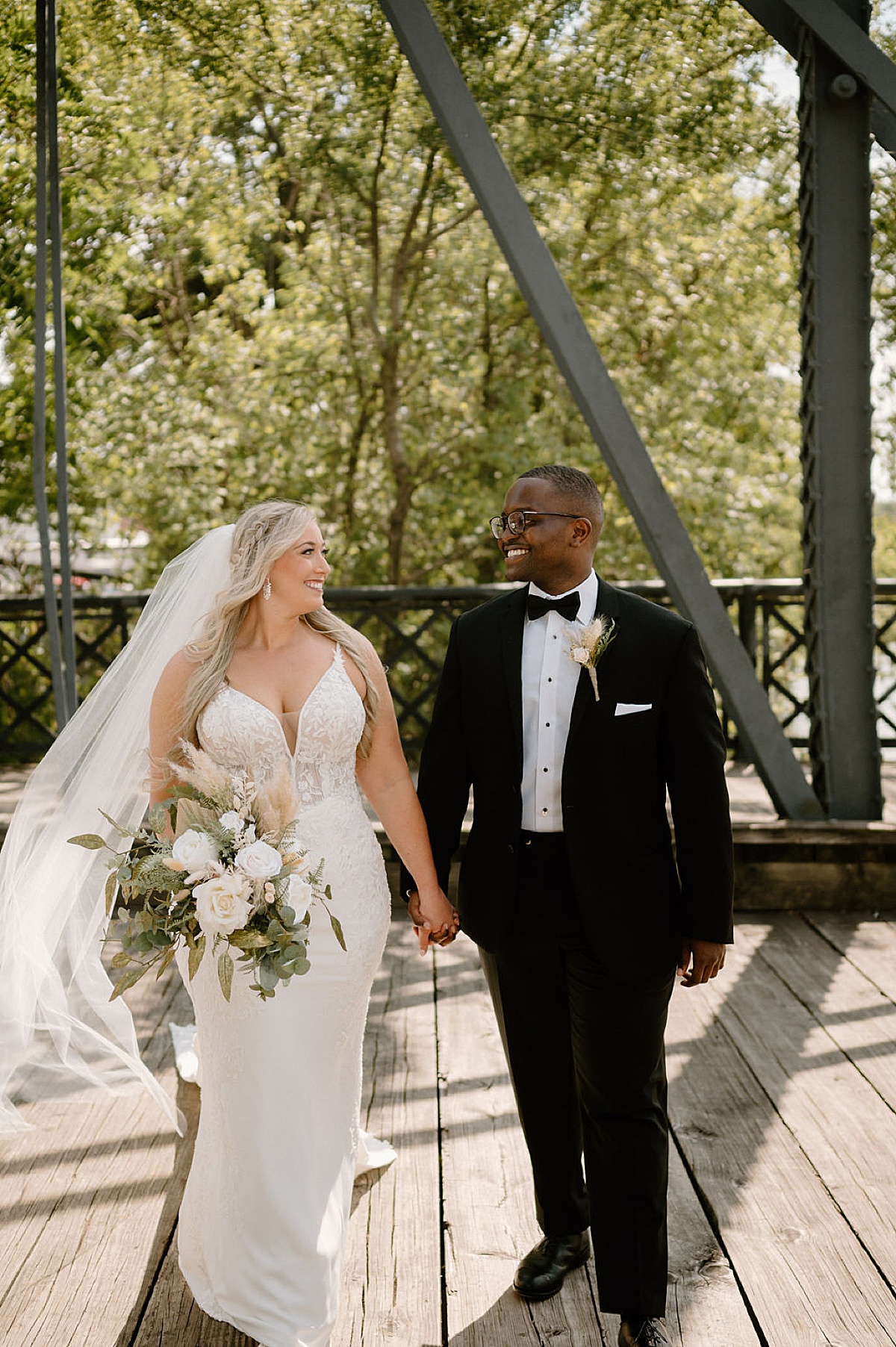 boho bride with pampas grass and eucalyptus rose bouquet poses with groom on sunny river bridge shot by midwest wedding photographer