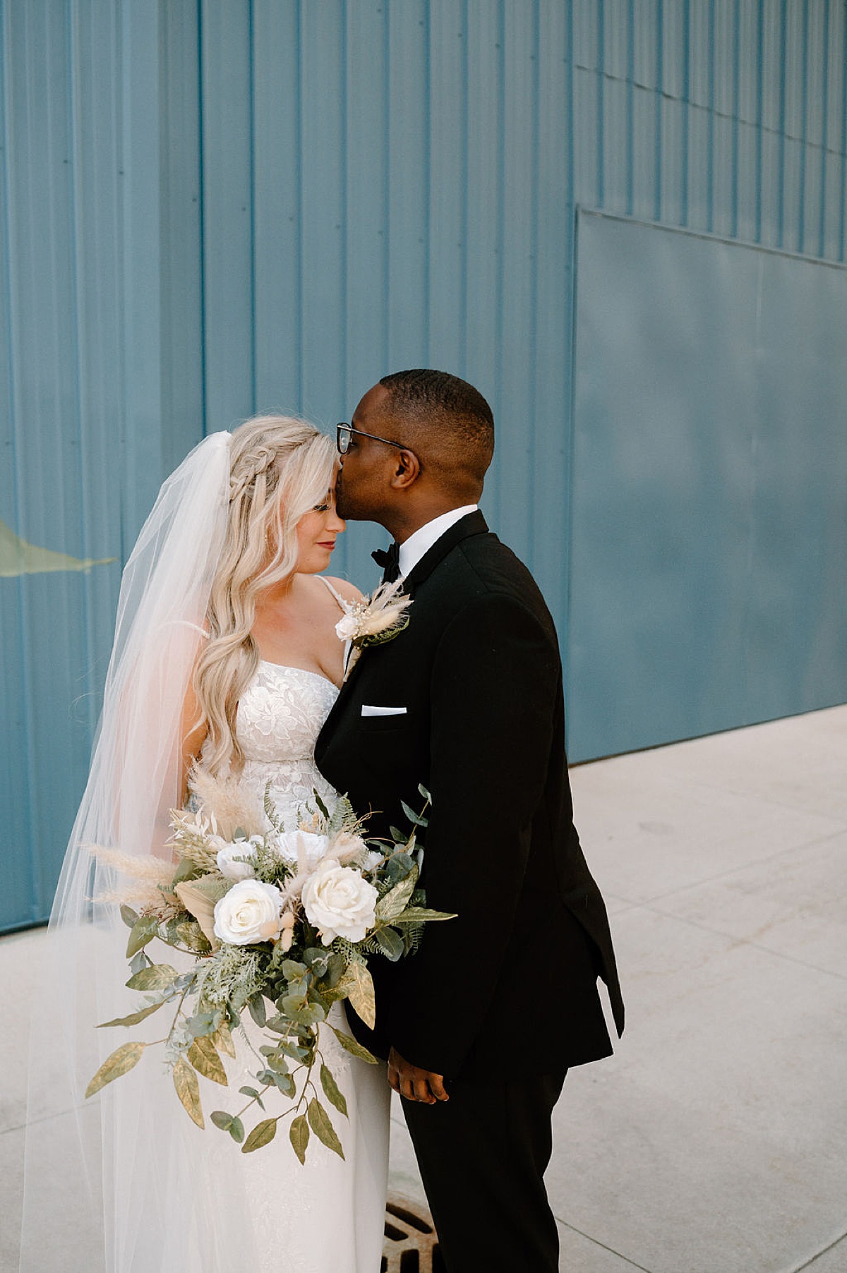 boho bride with braided hair and pampas grass and eucalyptus bouquet poses with groom in front of blue wall during photoshoot with midwest wedding photographer