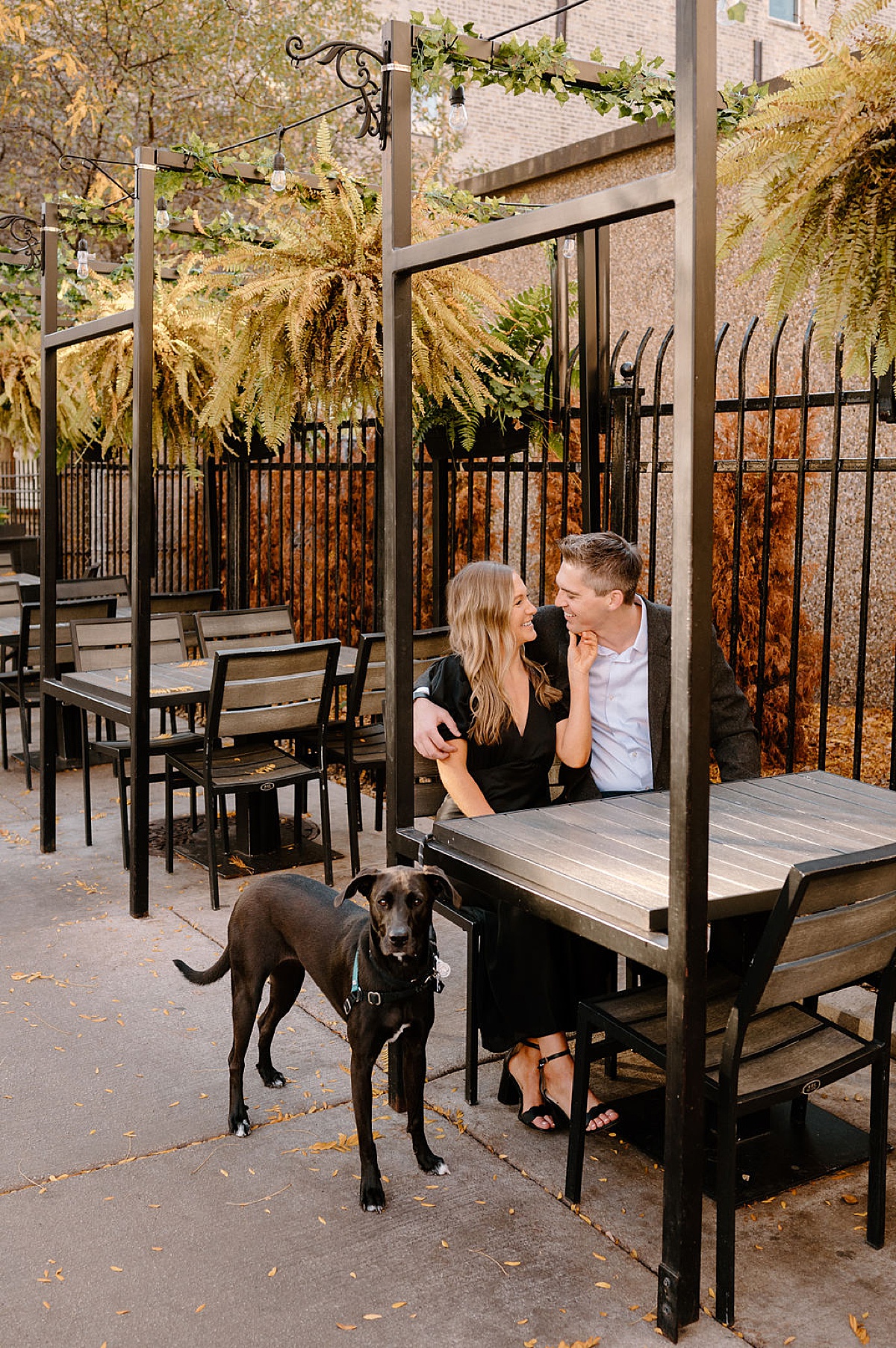 cute couple with family dog pose at outdoor cafe during engagement shoot with Chicago wedding photographer