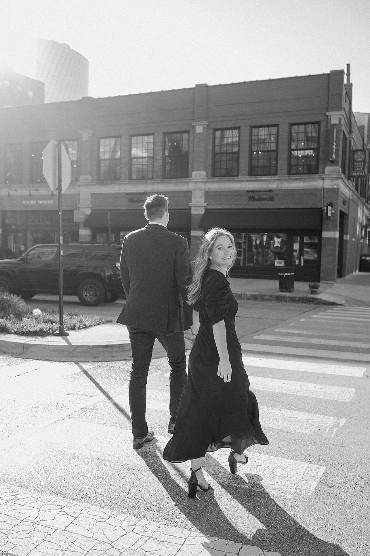 bride-to-be in classy black dress looks back over her shoulder while crossing a street with fiance shot by Chicago wedding photographer
