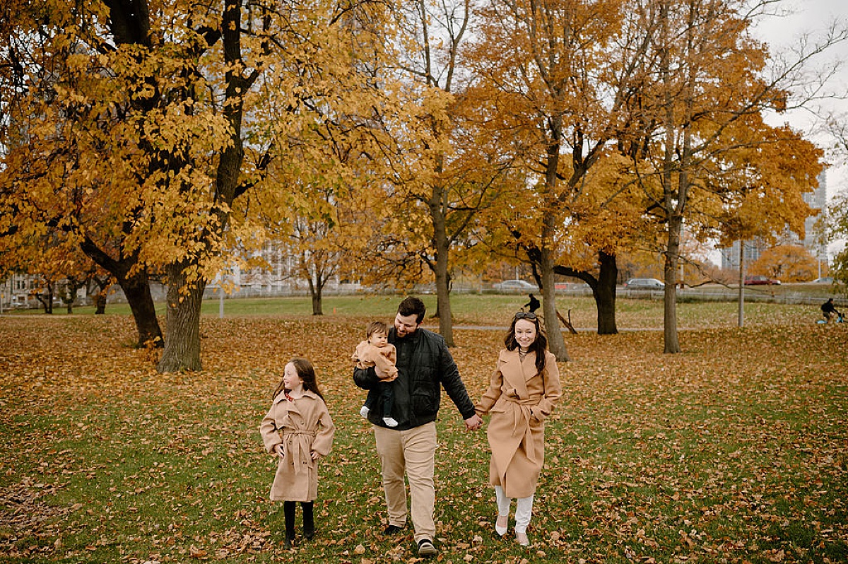 Cute family in camel overcoats walk hand in hand through fallen leaves during North Ave Beach Family Session