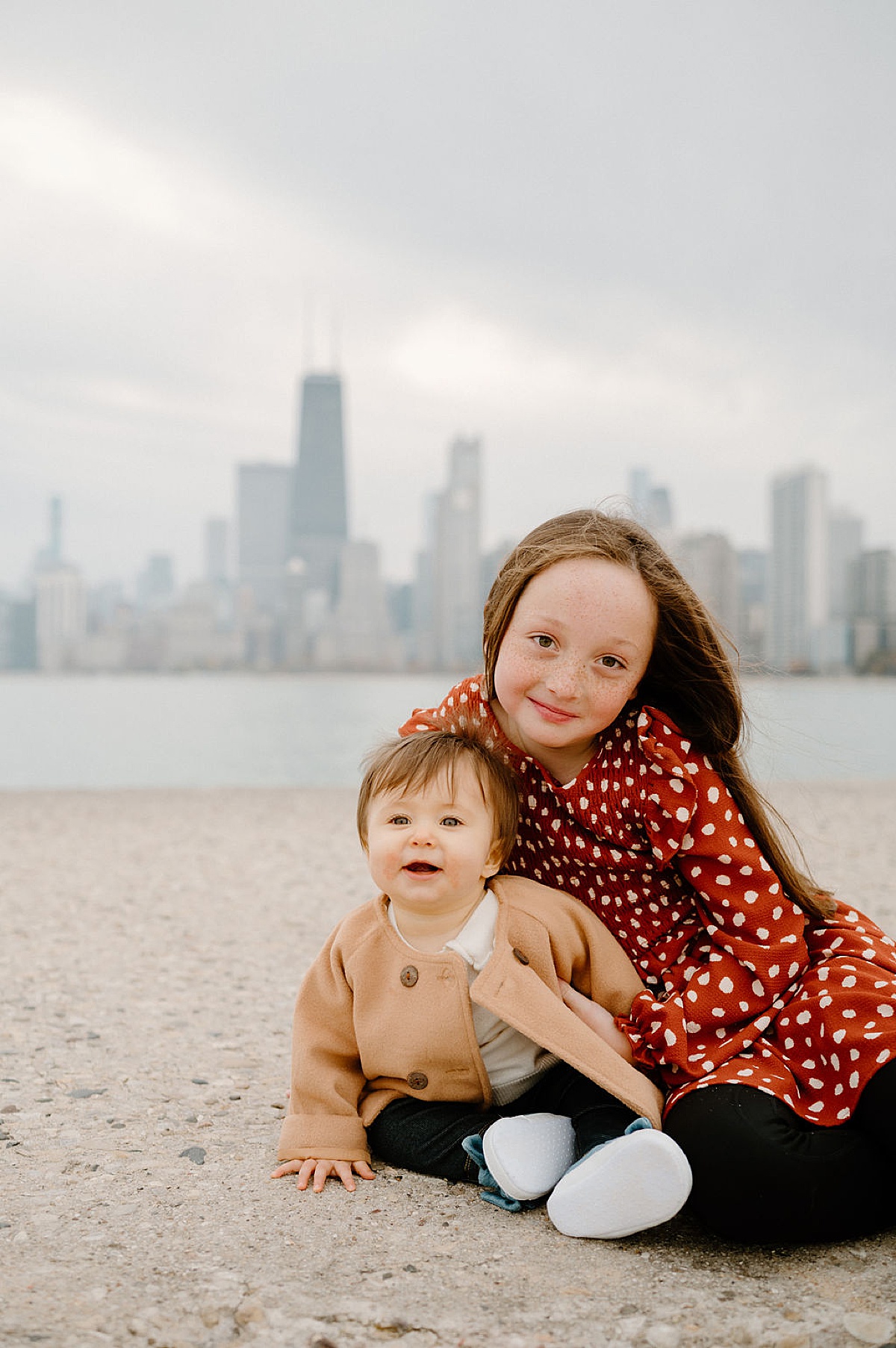 Little girl in polkadot dress poses with baby sister in front of Chicago skyline during family photos by Indigo Lace Collective