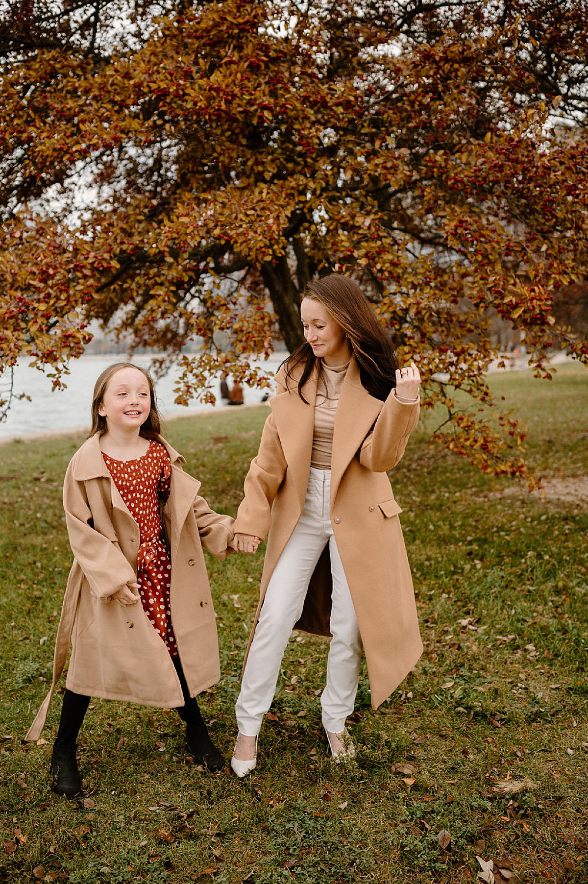 mom and preteen daughter poses in camel coats with fall leaves in the background during photoshoot with Chicago family photographer