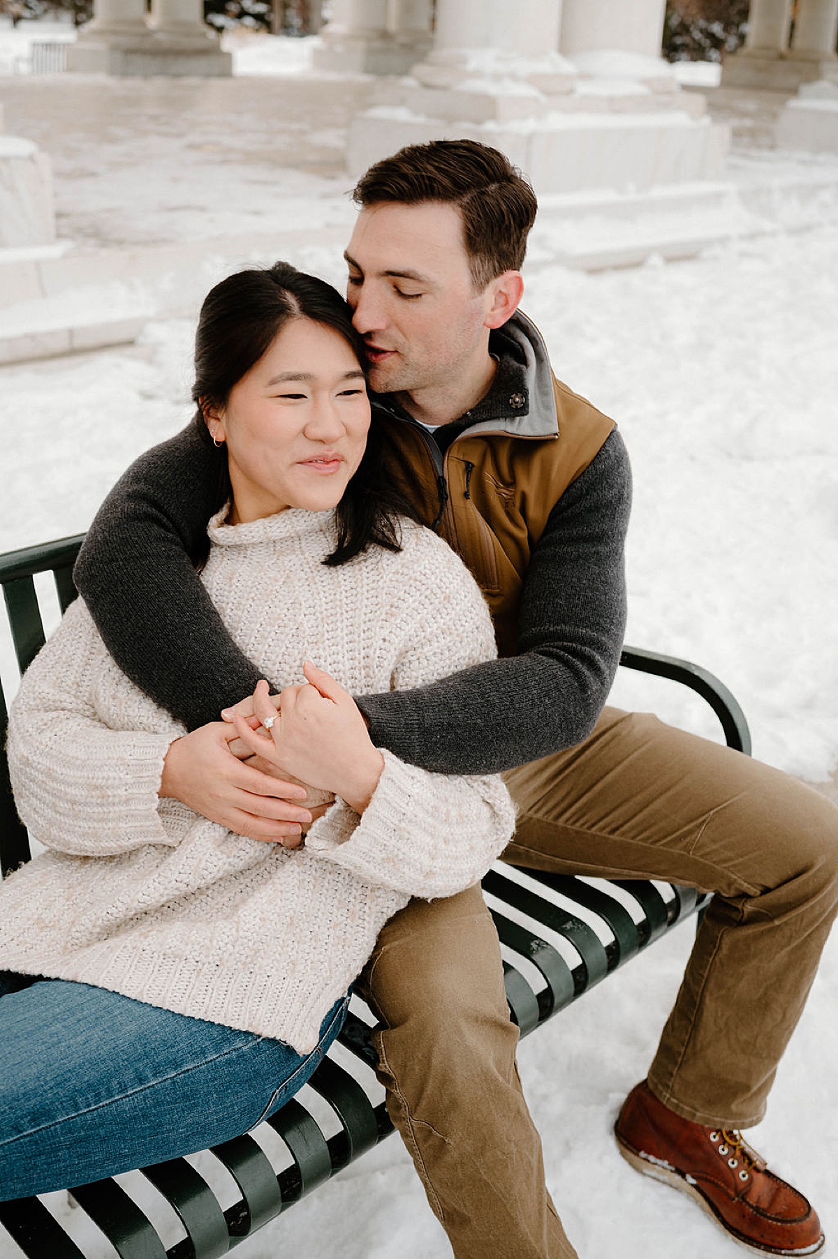woman in white sweater smiles as fiance kisses her forehead during snowy Cheesman Park engagement session