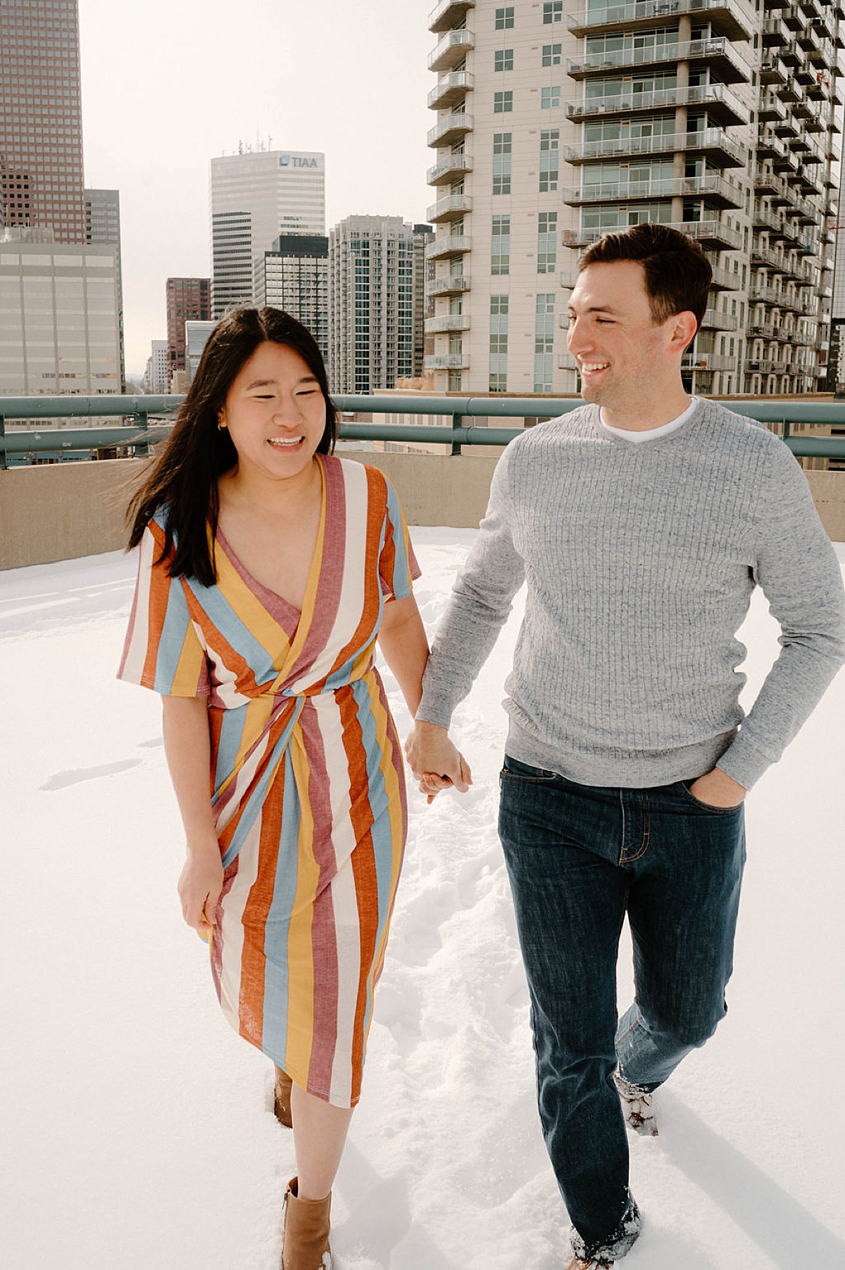 cute couple walk through snow on city rooftop during photoshoot with Denver wedding photographer