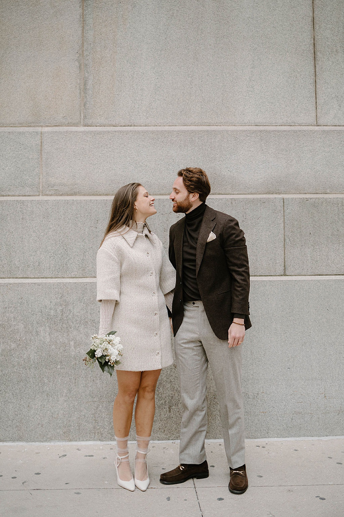 chic bride in mini length boucle coat  and groom in brown tweed jacket pose outside Winter City Hall Elopement