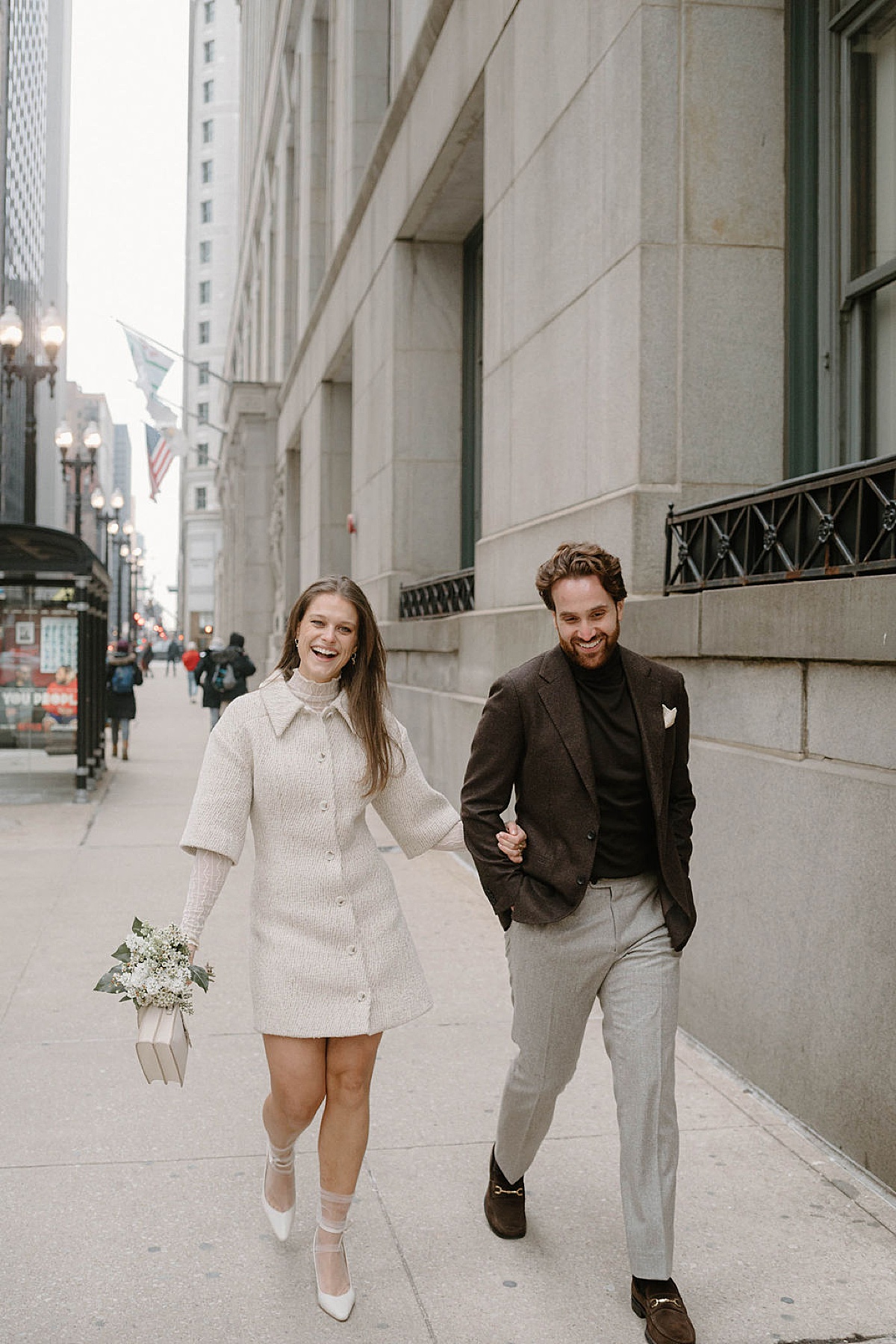 chic bride in boucle minidress and sheer socks and heels walks arm in arm with groom after Winter City Hall Elopement