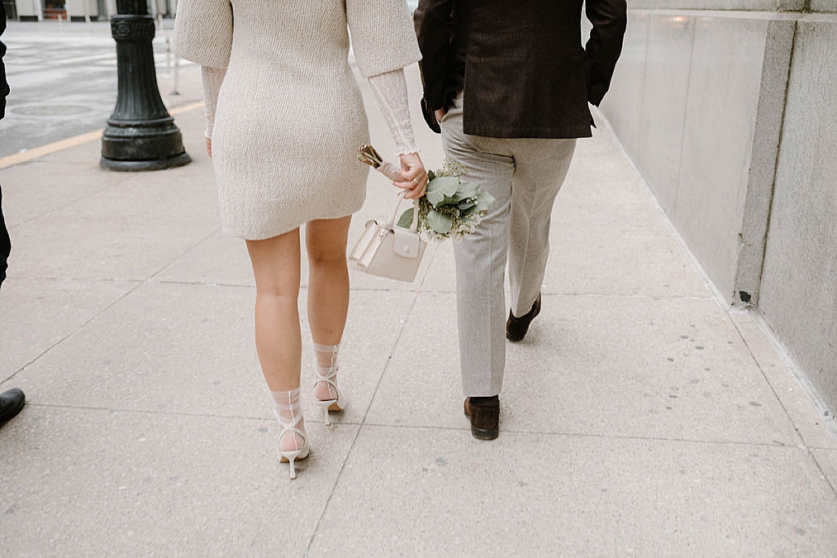 chic bride in boucle minidress coat and white heels walks with dapper groom through Chicago after Winter City Hall Elopement