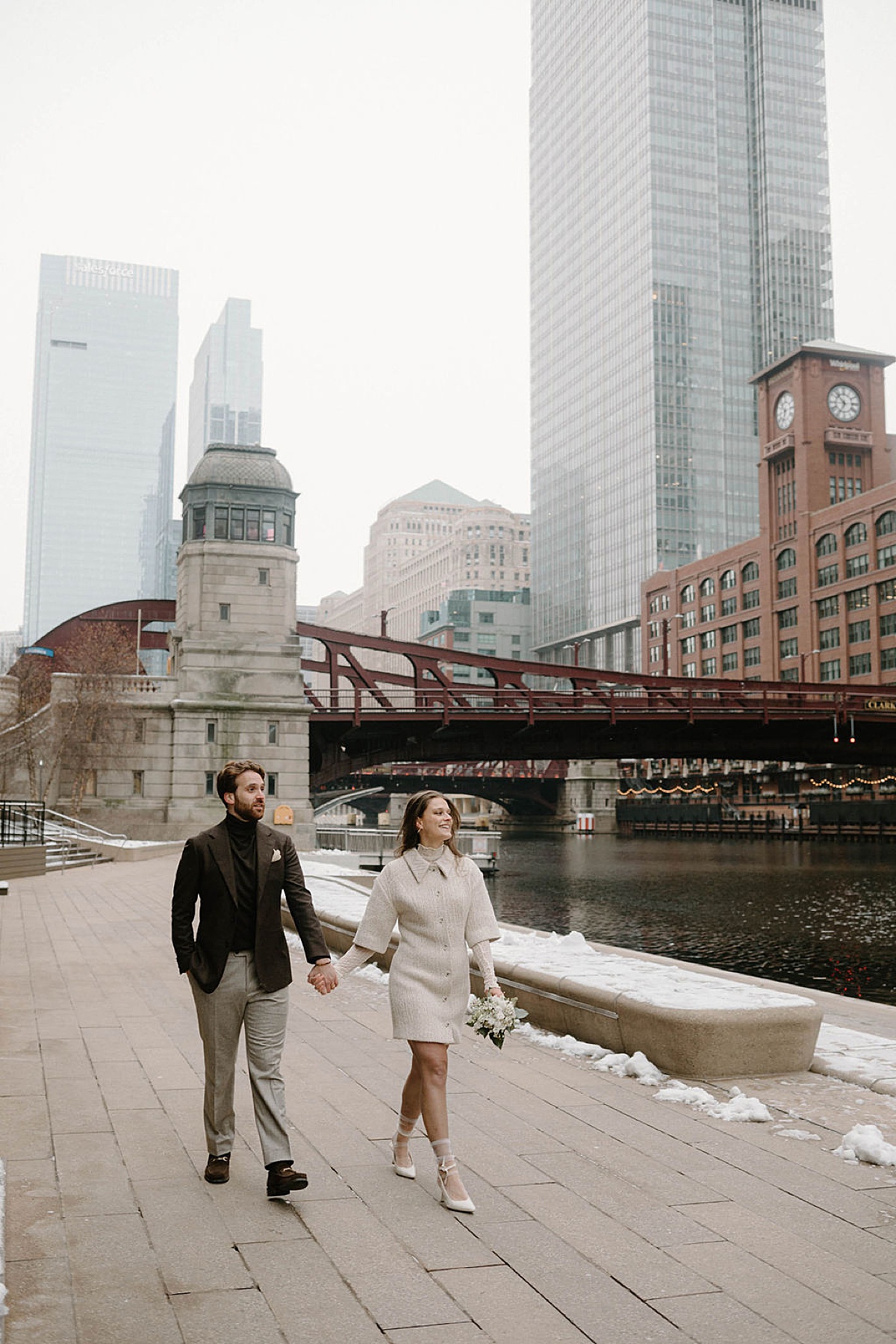 classy groom in tweed jacket and turtleneck walks with chic bride in minidress boucle coat down waterfront shot by Chicago wedding photographer