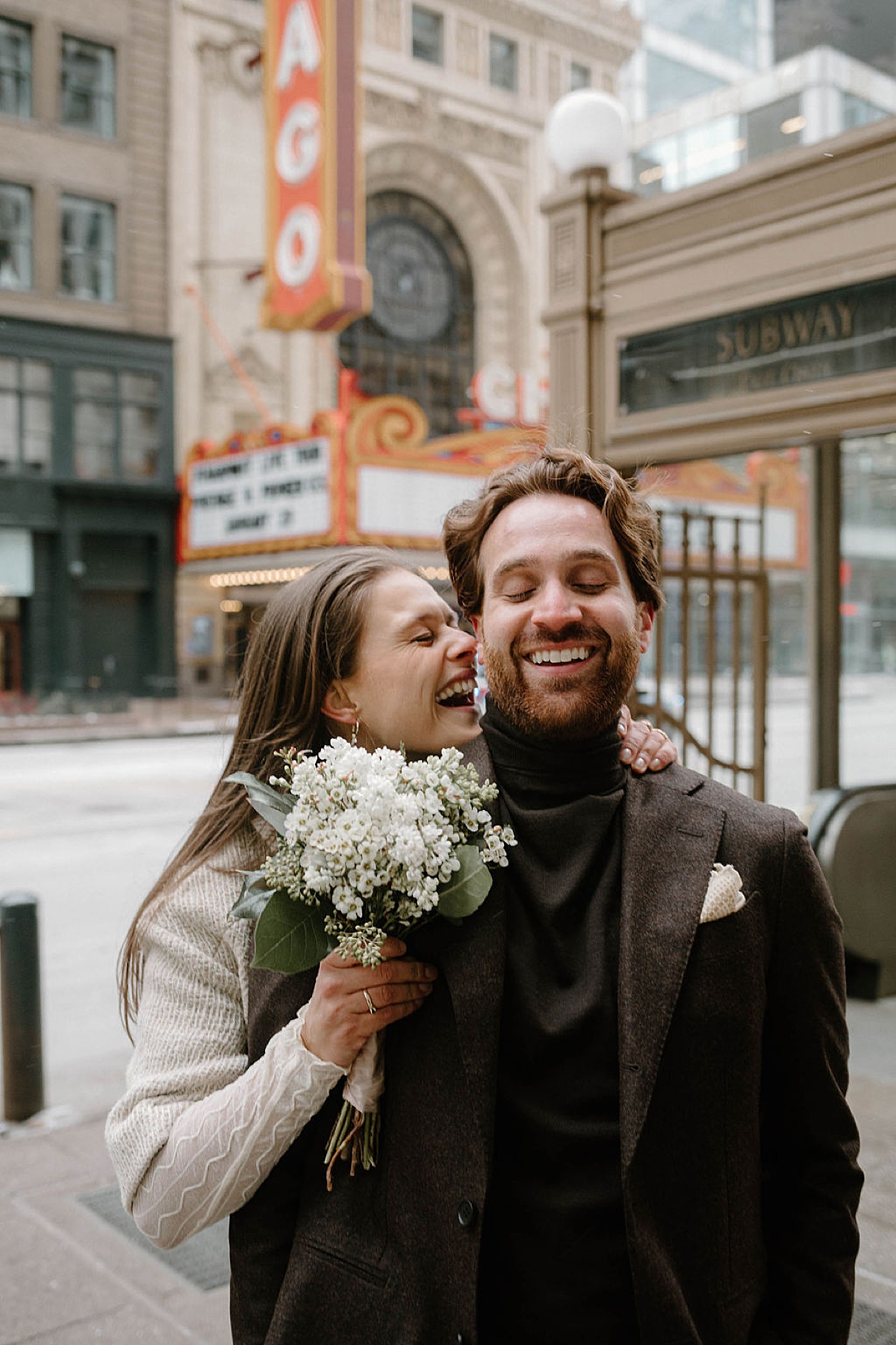 cute mod couple in coats with white flower bouquet pose in snowy city streets shot by Chicago wedding photographer