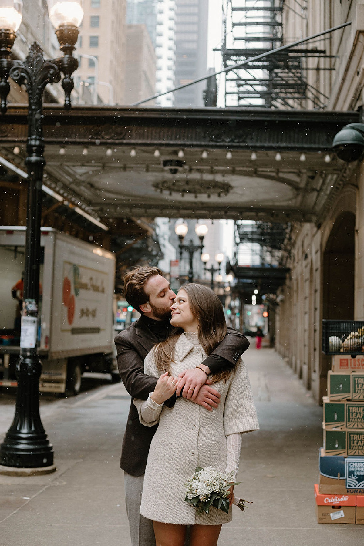 chic couple in mod 60s style coats embrace in snowy Chicago streets shot by Indigo Lace Collective