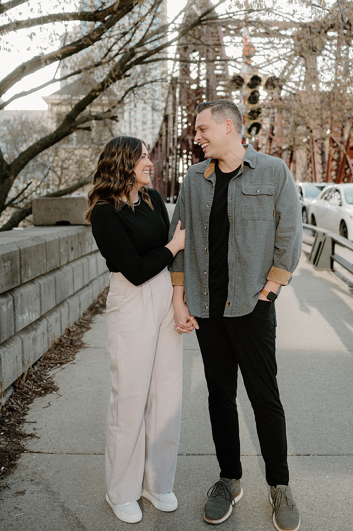 man and woman hold hands and walk across bridge during west loop city engagement shoot in Chicago