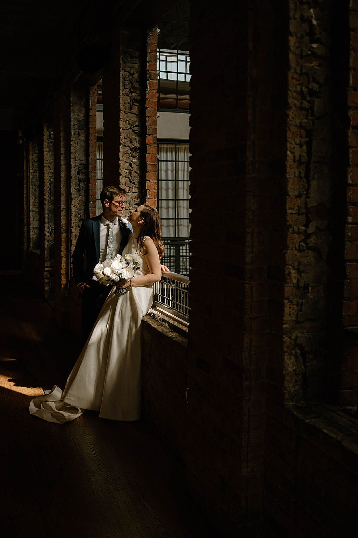 bride and groom pose in moody light from industrial window during gorgeous wedding shot by Indigo Lace Collective