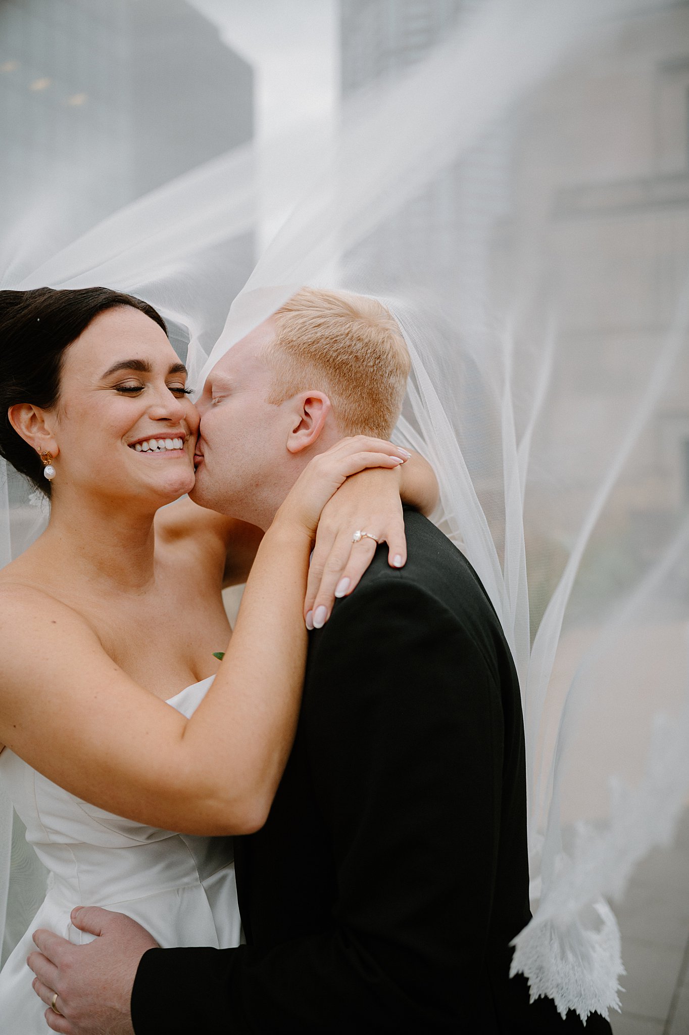groom kisses bride on the cheek as her veil blows in wind around them by Midwest wedding photographer