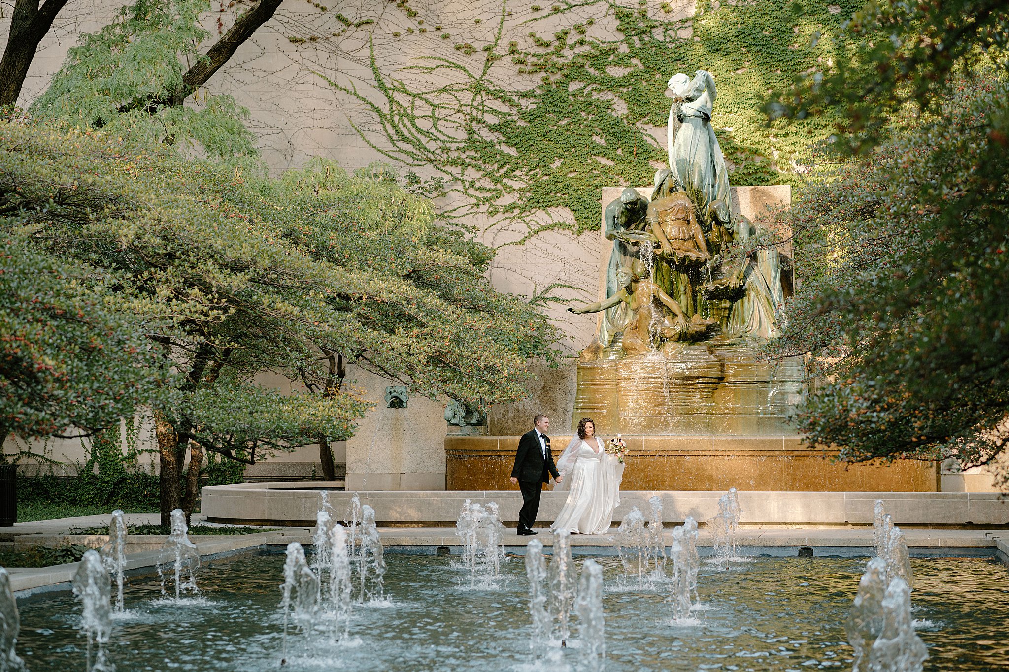 bride and groom hold hands as they walk past fountain by Indigo Lace Collective